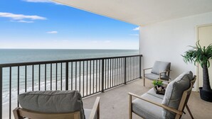 A balcony with a view of the ocean, two chairs, a small table with a plant, and a potted tree. - Regency Towers 1602 -Pool & Hot Tub Year-Round (Myrtle Beach)