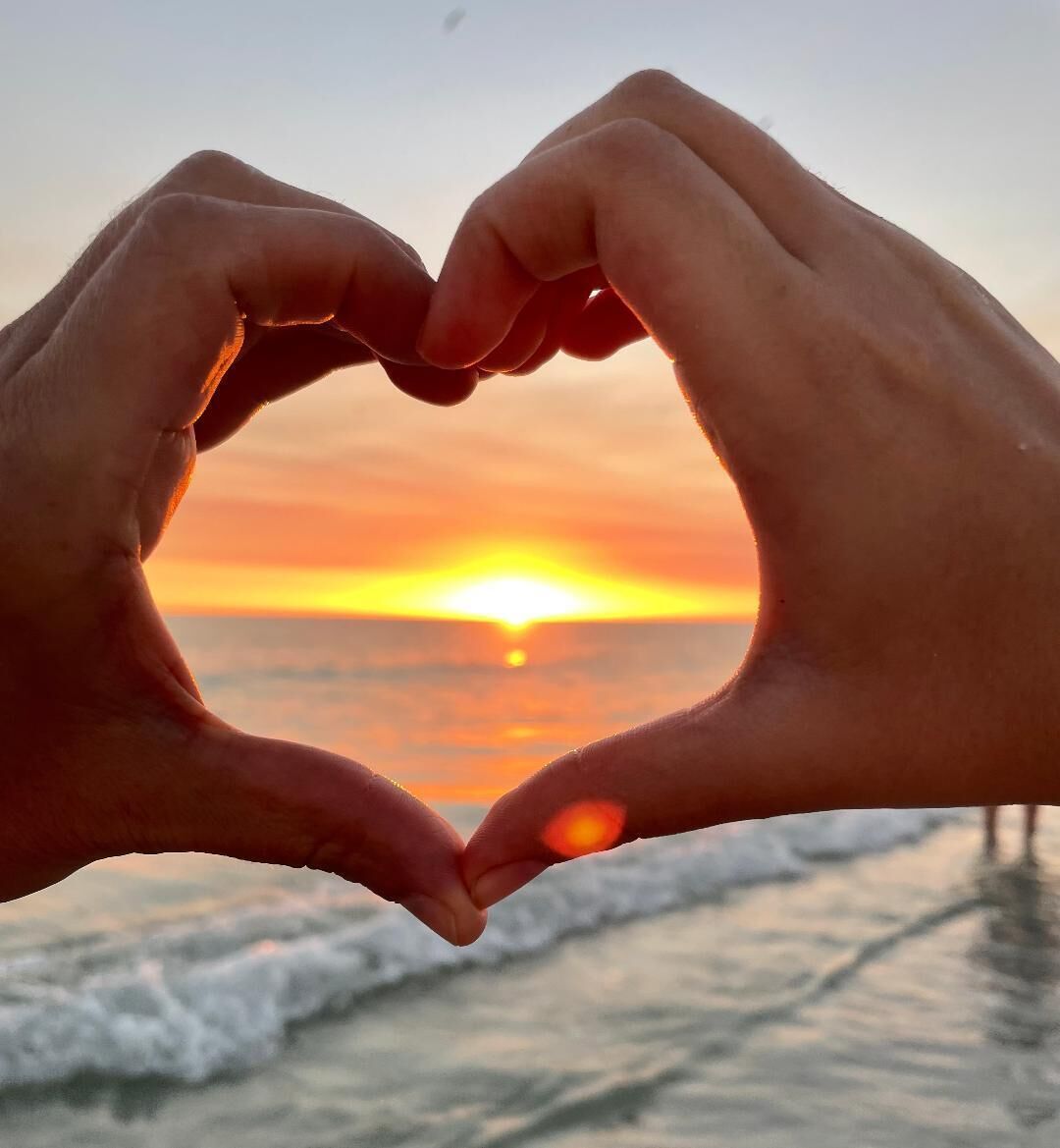 Des mains formant un cœur sur fond de coucher de soleil sur l’océan.