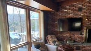 A living room with a brick fireplace, a television mounted on the wall, and a large window offering a view of the outdoors.