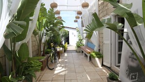 A narrow pathway flanked by large plants and a bicycle, leading to a bench and a door. - Rikas Alacati (Cesme)