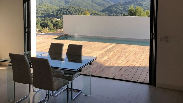 Una habitación moderna con una mesa y sillas de vidrio, techo de madera y un amplio ventanal corredero que da a una terraza con piscina y vistas a un paisaje montañoso.