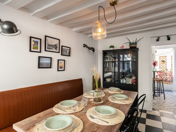 A dining area with a wooden table set for four, surrounded by a brown bench. There are framed pictures on the wall, a black cabinet with glass doors, and a hanging light fixture.