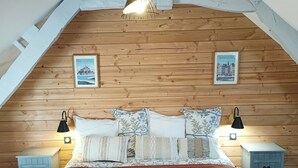 Un dormitorio con techo inclinado, paneles de madera en las paredes, una cama con varias almohadas, dos mesitas de noche con lámparas y dos cuadros enmarcados.