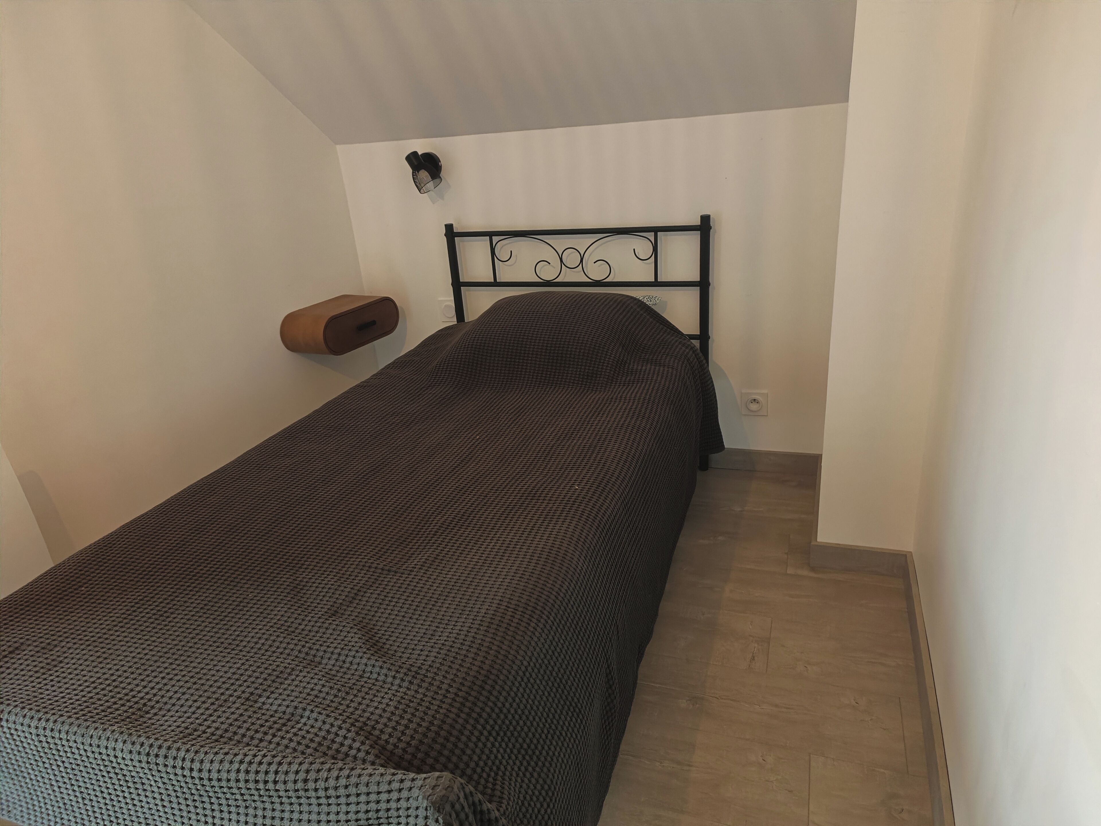 A single bed with a black metal headboard, a wooden nightstand, and a wall-mounted light fixture.