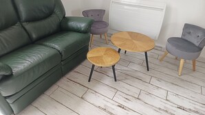 Un sofá de cuero verde, dos mesas redondas de madera y dos sillas en una habitación con un radiador blanco y suelo de baldosas.
