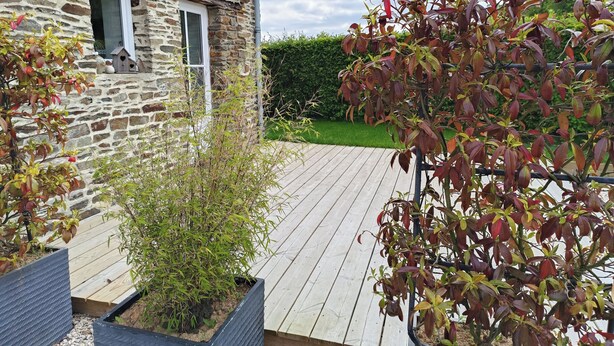 Un edificio de piedra con una terraza de madera, un pequeño jardín y un seto al fondo.