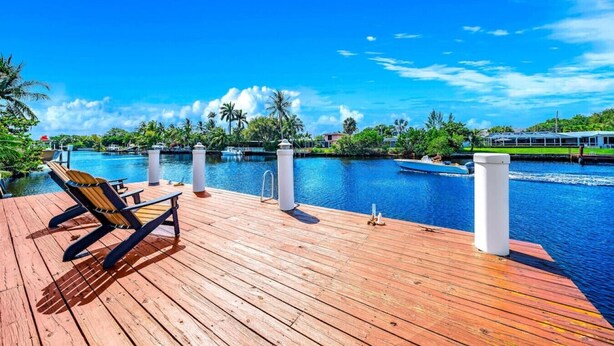 Una terraza de madera con sillones de descanso con vistas a un río tranquilo, rodeada de exuberante vegetación y un cielo azul despejado.