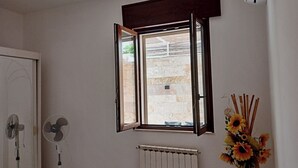 Ein Zimmer mit Fenster, Heizkörper, Standventilator und Blumen in einer Vase.