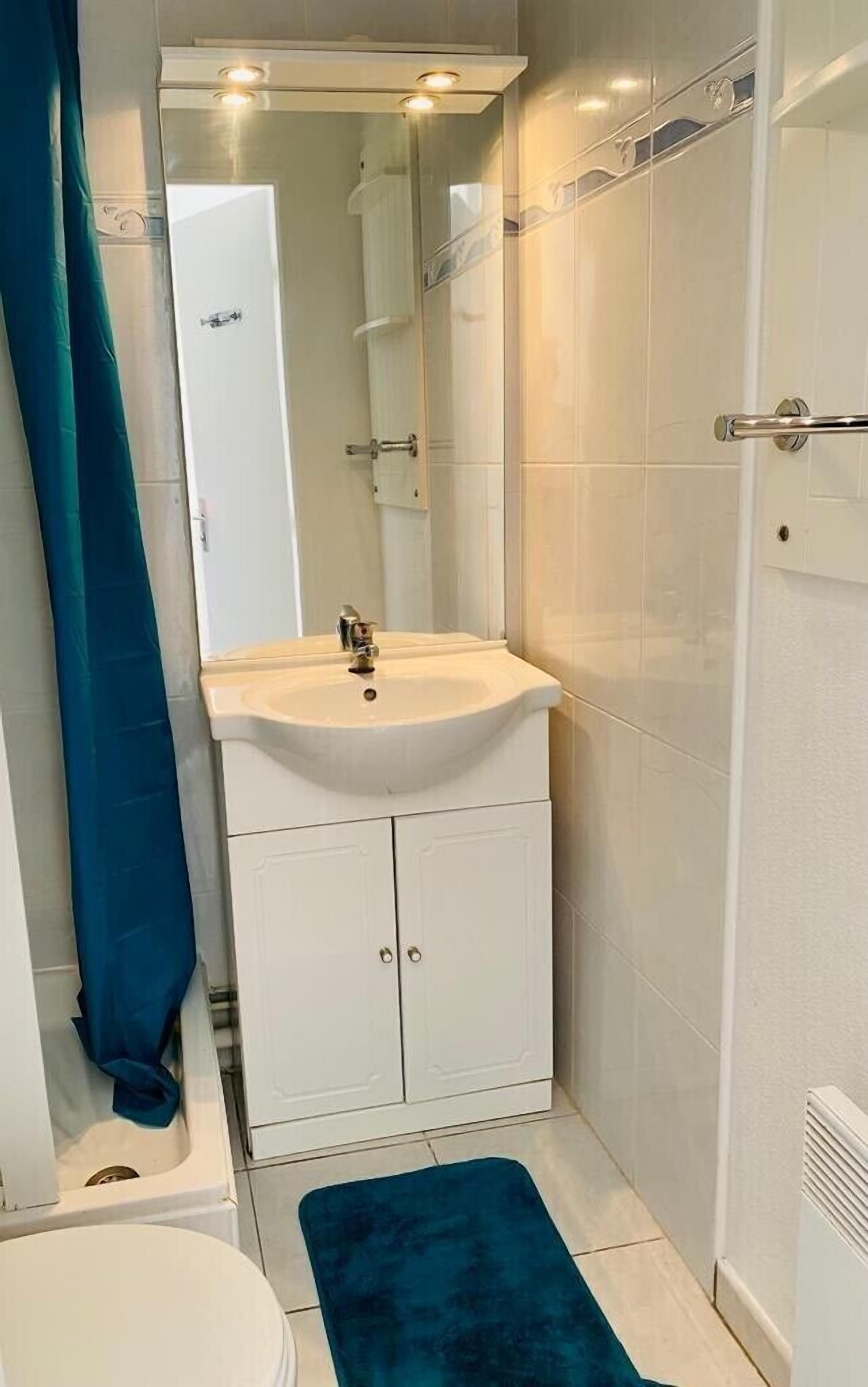Une petite salle de bain avec un lavabo blanc, un miroir au-dessus, un espace douche et un tapis de bain bleu.