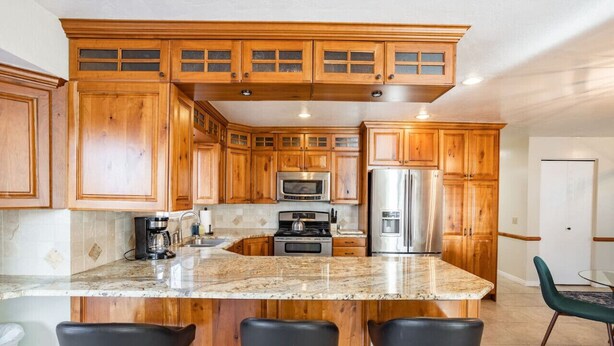 A kitchen with wooden cabinets, a granite countertop, stainless steel appliances, and black bar stools. - Spacious 3BR Extended Stay: Airport, Downtown, Ski (North Salt Lake)