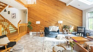 A spacious living room with a high ceiling, wooden paneling, a staircase, a fireplace, and a large window. - Modern, Magical Woodsy Escape 1 Hour From NYC (Putnam Valley)