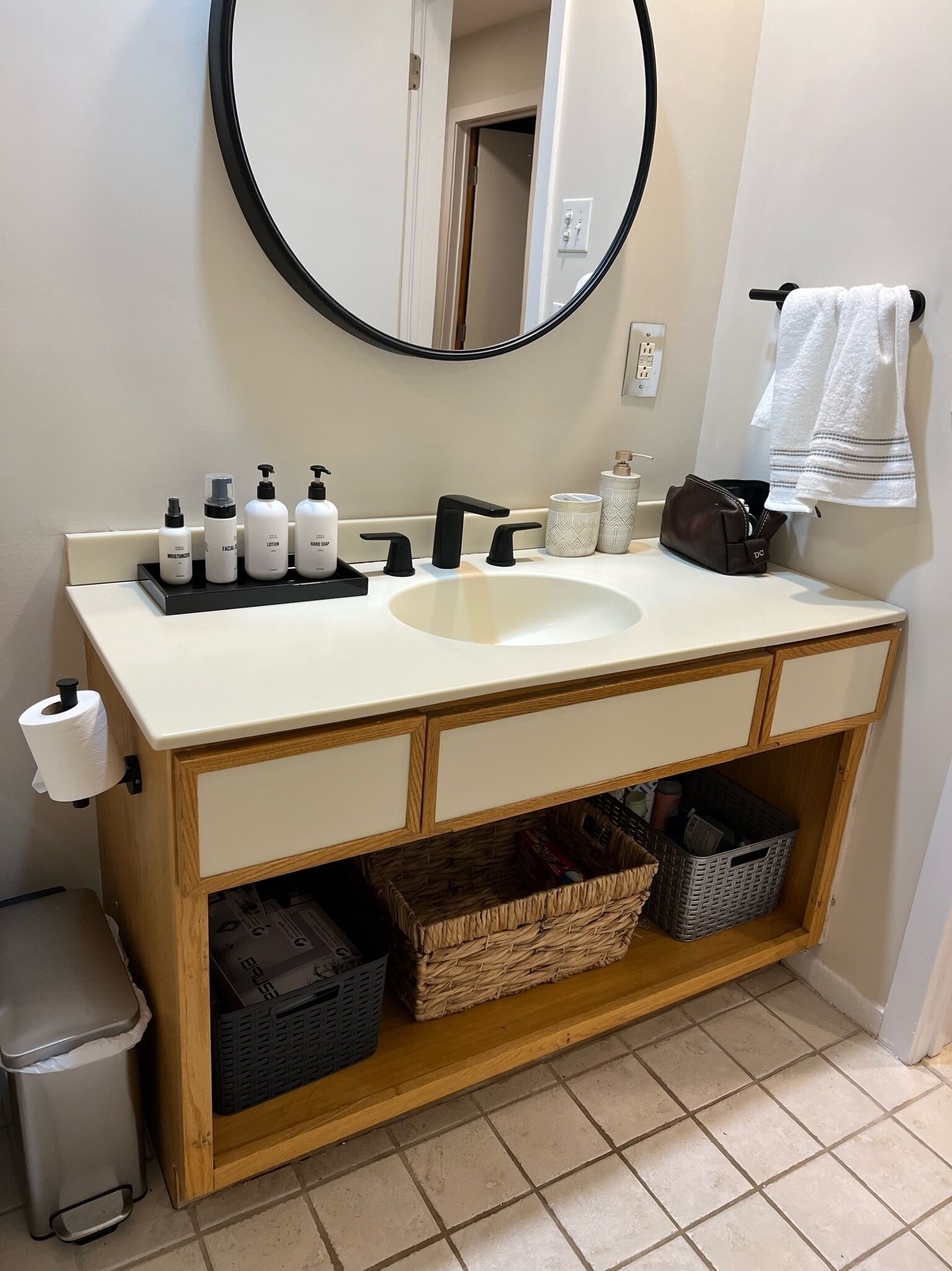 Un baño con un lavabo blanco, grifo negro y un espejo encima. Hay artículos de aseo en el lavabo, un cubo de basura y un perchero con una toalla.