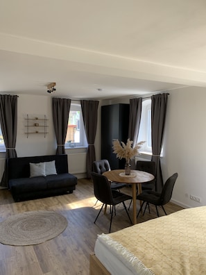A modern room with a bed, a sofa, a dining table with chairs, a small rug, and a window with curtains. - Vacation apartment I Beck-Bräu near Bamberg (Lisberg)