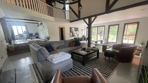 A spacious living room with a large sectional sofa, a coffee table, and a leather armchair. There is a staircase leading to an upper level, and large windows providing natural light. - Modern Rustic 5 Bedroom Home In the Foothills of the Catskills Mountains. 5 ⭐️ (Otego)