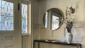A hallway with a white door, a round mirror, a white vase with flowers, a woven basket, and a shelf with books. - Modern Rustic 5 Bedroom Home In the Foothills of the Catskills Mountains. 5 ⭐️ (Otego)
