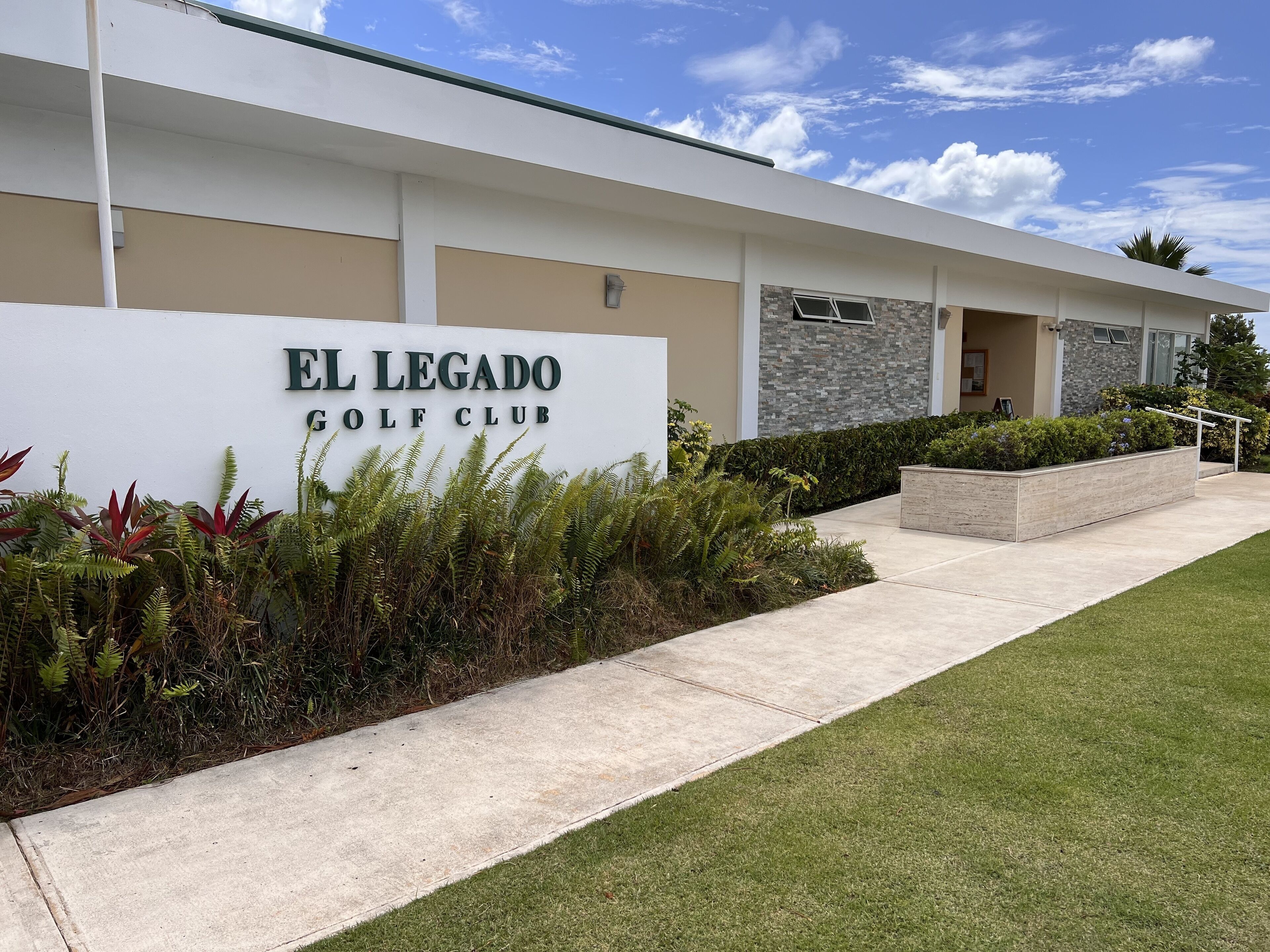 The exterior of El Legado Golf Club with a sign, a paved walkway, and landscaped greenery.