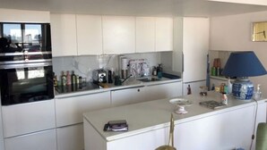 Une cuisine moderne avec des armoires blanches, un four encastré, une machine à café et un petit îlot central agrémenté d’une lampe.