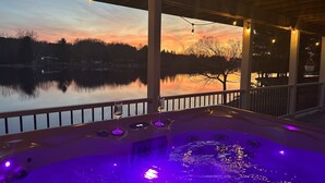 Banheira de hidromassagem com vista para um pôr do sol sobre um lago, cercada por um deck com guarda-corpos.