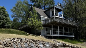 Uma casa de dois andares com varanda, cercada por um muro de pedra e uma área arenosa com cadeiras Adirondack.