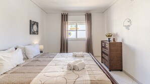 A hotel room with a bed, a dresser, a window with curtains, a wall-mounted light fixture, and a wall decoration. - #075 T1 Solar by Home Holidays (Albufeira)