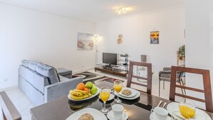 A modern living room with a sofa, dining table with breakfast, TV, and artwork on the wall. - #075 T1 Solar by Home Holidays (Albufeira)