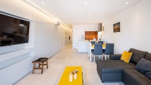 Uma sala de estar moderna com televisão de tela plana, um sofá, uma área de jantar e uma cozinha ao fundo.