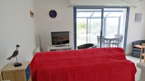 A room with a bed covered in a red blanket, a television on a white cabinet, a wall clock, and a window with a view of the outside. - Bright, sea-view apartment with direct beach access (Roscoff)