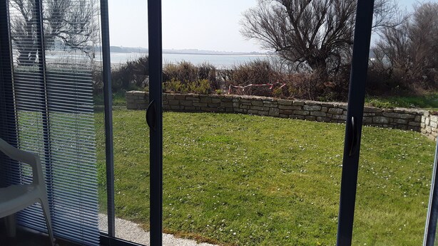 A view through a glass door of a grassy outdoor area with a stone wall and a tree. - Bright, sea-view apartment with direct beach access (Roscoff)