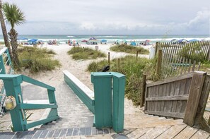 A beach with a wooden walkway leading to the shore, surrounded by beach umbrellas and people.