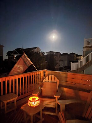 A wooden deck with a chair and a table, a lit lantern, and a view of houses under a night sky with a moon.