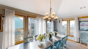 A dining area with a table set for four, surrounded by chairs, a chandelier, and large windows with curtains. - Semi Oceanfront 5 bedroom dog friendly cottage 10 feet to beach access (Corolla)