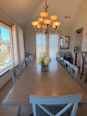 Een eetzaal met een lange tafel, stoelen, een kroonluchter en een vaas met bloemen.