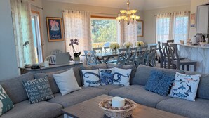 A living room with a sectional sofa, a dining area with a table and chairs, a kitchen area, and a large window with curtains. - Semi Oceanfront 5 bedroom dog friendly cottage 10 feet to beach access (Corolla)
