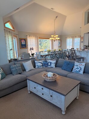 A living room with a sectional sofa, a dining area with a table and chairs, a kitchen area, and a large window with curtains.