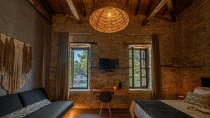 Une pièce rustique aux murs en pierre apparentes, avec un plafond en bois, un luminaire en fibres naturelles, un lit, un canapé, un bureau avec une chaise et un téléviseur fixé au mur.