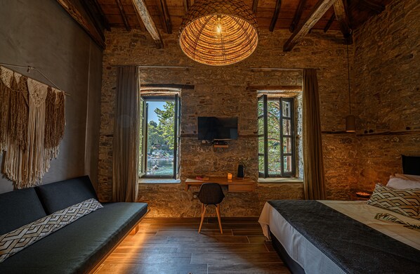 Una habitación rústica con paredes de piedra vista, techo de madera, lámpara de fibra natural, cama, sofá, escritorio con silla y televisor en la pared.