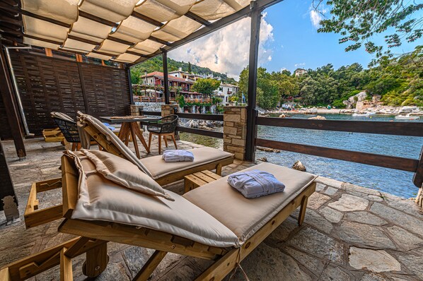 A covered outdoor seating area with wooden loungers and stone flooring, overlooking a body of water and a hillside with buildings.