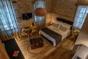 A bedroom with a stone wall, a bed with a headboard, a television, a bedside table, a suitcase, and a window with curtains.