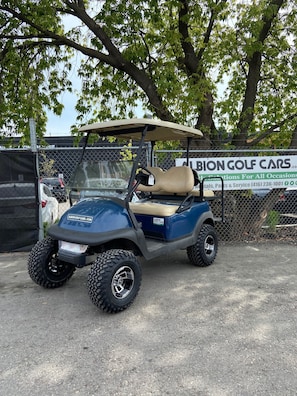 Un golf cart parcheggiato in un parcheggio, con una recinzione e alberi sullo sfondo.