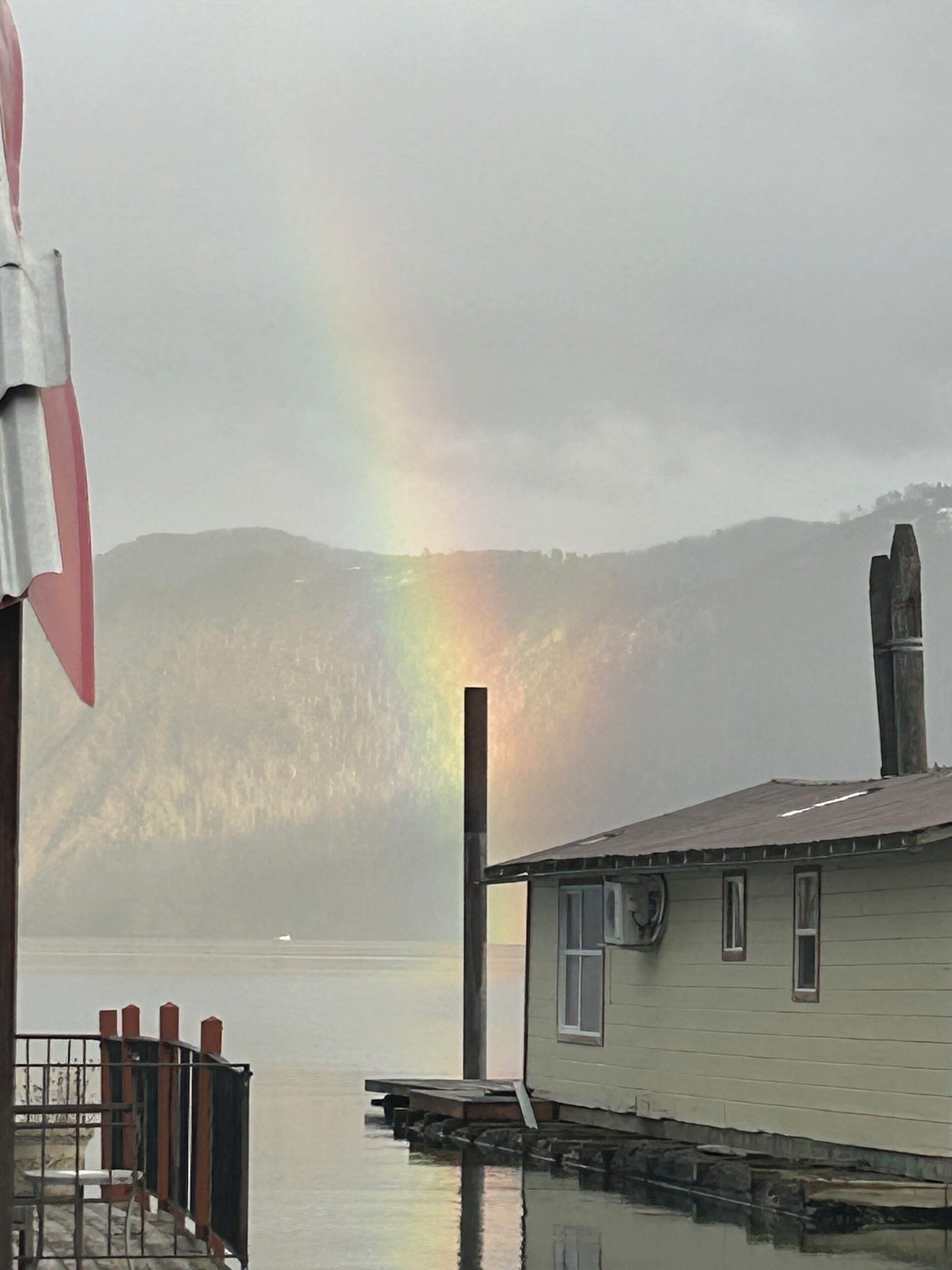 Une maison au bord de l’eau, avec un arc-en-ciel en arrière-plan.