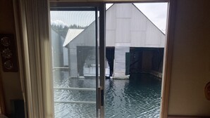 Een kamer met een grote glazen schuindeur die uitkomt op een houten terras boven water, een bed met een beige deken en een kamerkleed met een patroon.