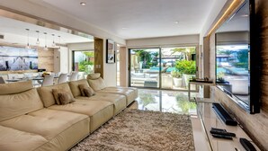 Ein geräumiges Wohnzimmer mit einem großen, beige gefärbten Sofa, einem Glastisch und einem an der Wand montierten Flachbildfernseher.