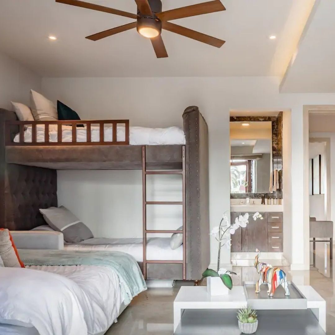 Un dormitorio moderno con literas, ventilador de techo, una kitchenette pequeña y una mesa de centro con objetos decorativos.