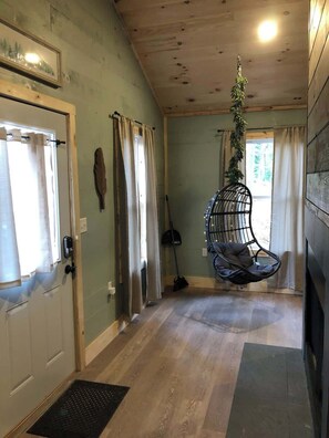 A room with a hanging chair, a door with curtains, a wall-mounted coat rack, and a window with a view of greenery. - Butterfly Ridge at Avenwood Cabins (New Straitsville)