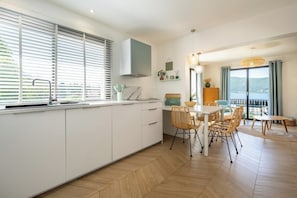 A modern kitchen with white cabinets, a dining area with wicker chairs, and a view of the sea.