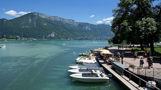 A lakeside view with boats docked, a promenade, and mountains in the background.