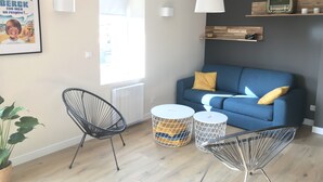 Een moderne woonkamer met een blauwe bank, twee zwarte draadframe stoelen, een witte lamp en een ingelijst poster aan de muur.