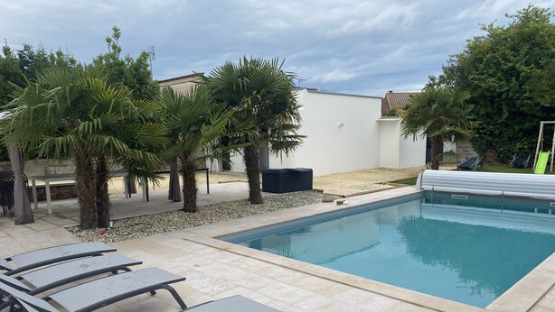 A swimming pool with lounge chairs, palm trees, and a white wall in the background. - Villa Royan 10 persons with private heated pool 650m from the beach (Royan)