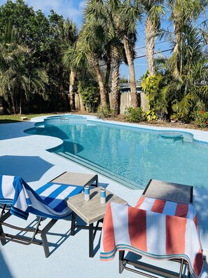 Área de piscina com espreguiçadeiras e mesa, cercada por palmeiras e um muro de madeira.