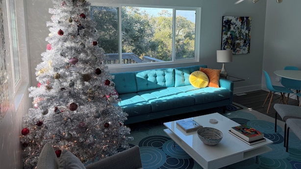 A modern living room with a large window, a teal sofa, a white coffee table, and a Christmas tree.